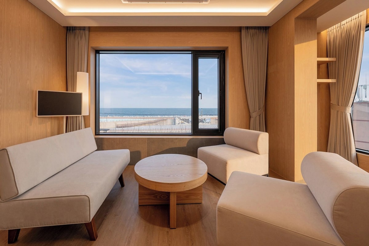 A hotel room with a large window, a sofa, a chair, a small table, and a TV mounted on the wall.