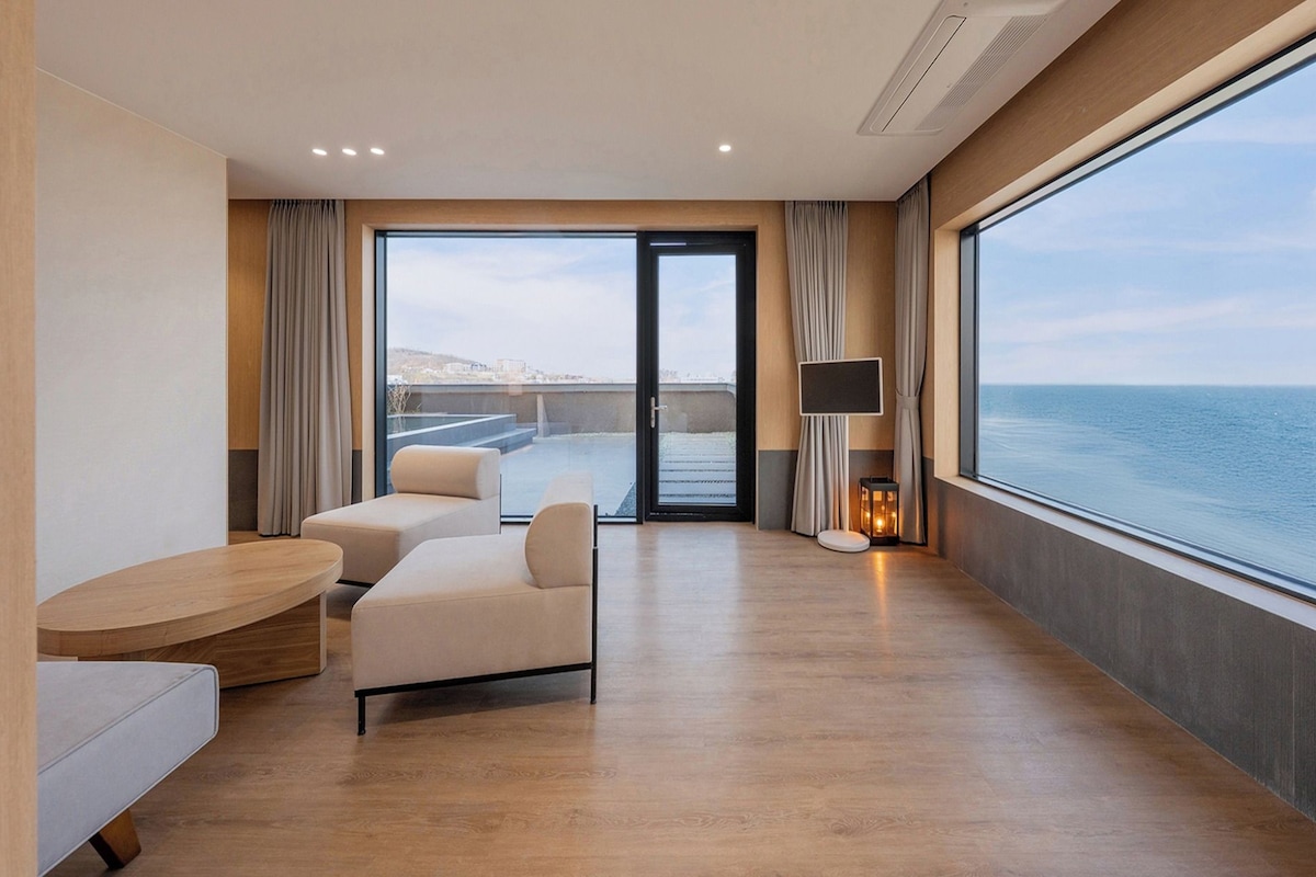 A modern hotel room with a balcony, a seating area with a table, and a large window offering a view of the sea.