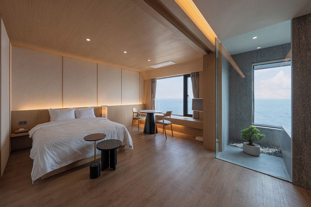 A modern hotel room with a large bed, a small round table, and a view of the sea.