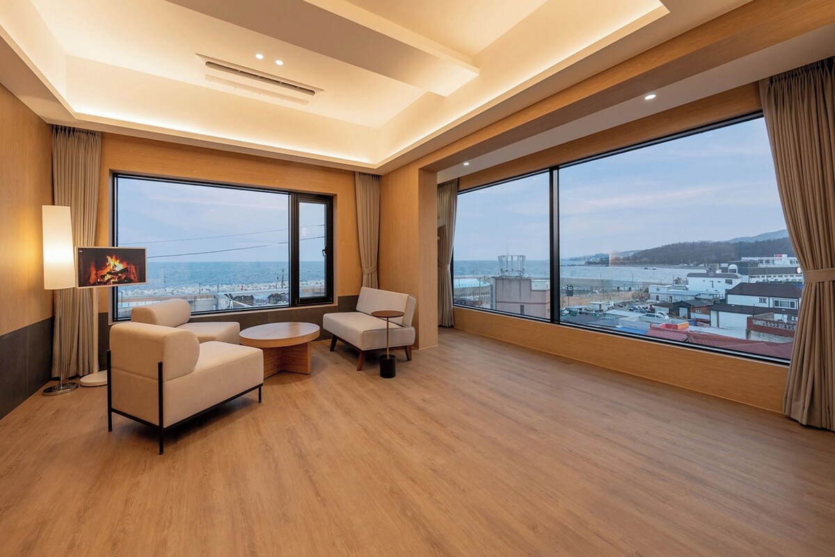 A hotel room with a large window offering a view of the sea and cityscape, a sofa, a small round table, and a flat-screen TV mounted on the wall.