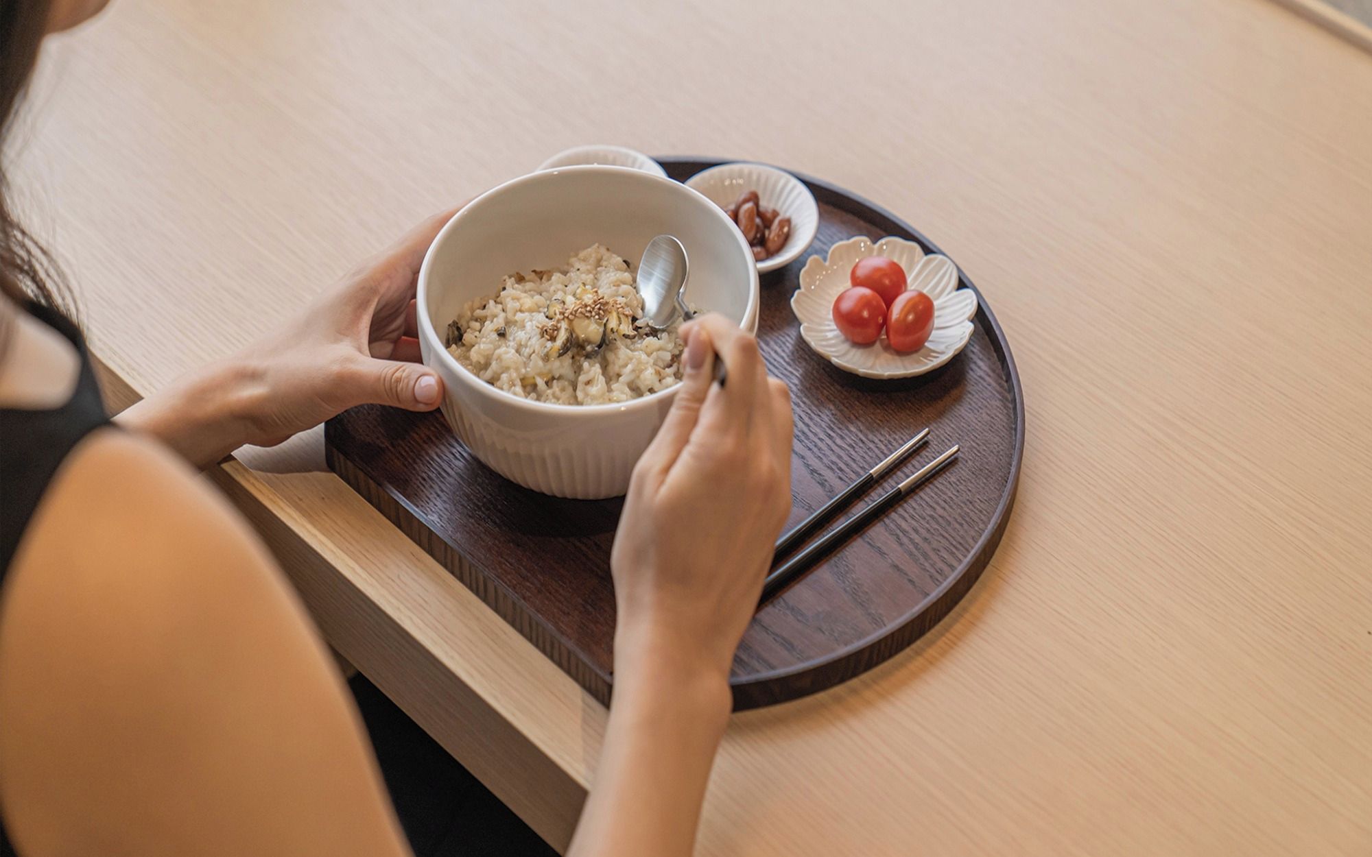 A person eating rice with a spoon, a small plate of cherry tomatoes, and chopsticks on a wooden tray.