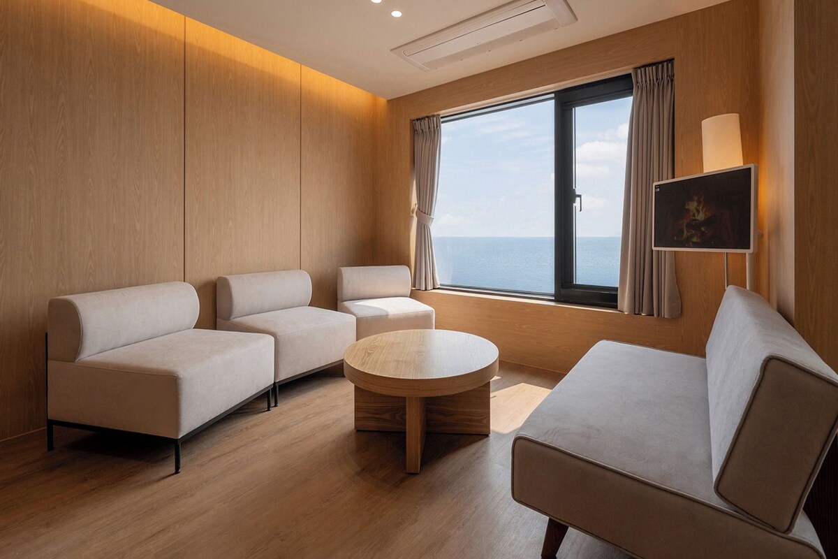 A modern hotel room with a sofa set, a round coffee table, and a large window offering a view of the sea.