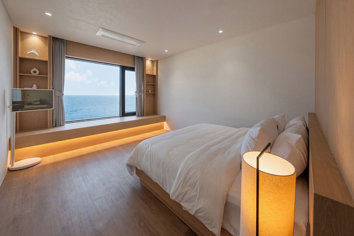 A bedroom with a large bed, a bedside lamp, a TV on a shelf, and a view of the sea.