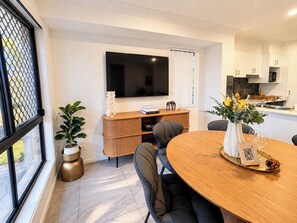 Un espace repas moderne avec une table ronde, des chaises, un téléviseur fixé au mur et une cuisine en arrière-plan.