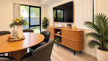 A modern dining area with a round wooden table, leather chairs, a white vase with flowers, and a built-in TV on a wooden cabinet.