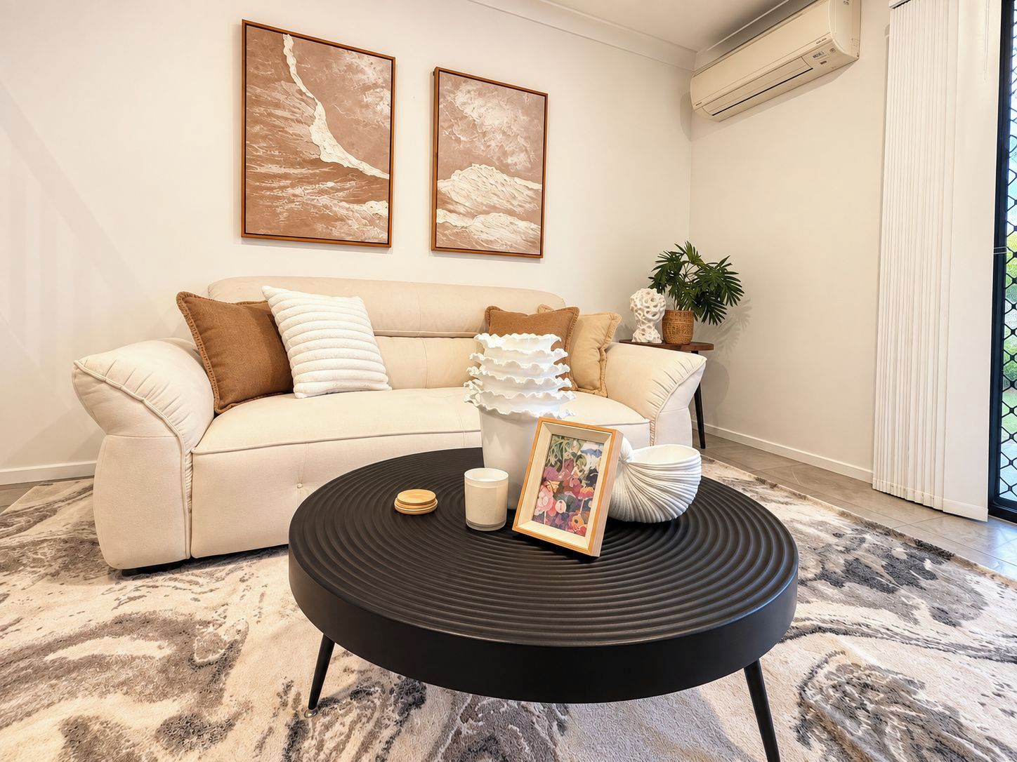 A living room with a beige sofa, two framed artworks, a black coffee table with a picture frame, and a potted plant.