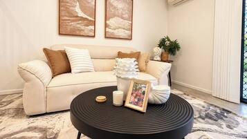 A living room with a beige sofa, two framed artworks, a black coffee table with a picture frame, and a potted plant.