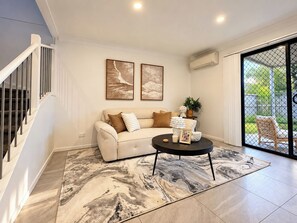 A modern living room with a staircase, a sofa, a coffee table, and a large window with a view. - Boondall 3BR Family Home | Long Stay Deals (Boondall)