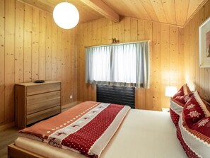 Een kamer met houten vloeren, een bed, een dressoir, een raam met gordijnen en een hanglamp.