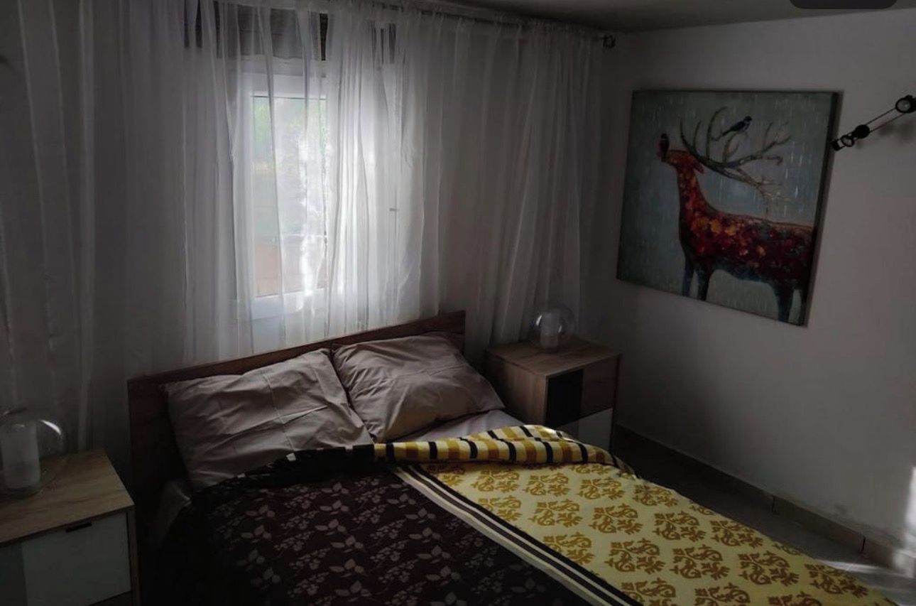 A bedroom with a bed, a nightstand, a window with sheer curtains, and a painting of a deer on the wall.