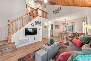 A spacious living area with a wooden staircase, a flat-screen TV mounted on the wall, a dining area with wooden chairs, and a large window. - Little Pinecone Retreat close to Rec Center (Truckee)