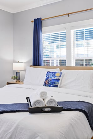 Ein Hotelzimmer mit einem Bett, einem Nachttisch mit Lampe, einem Fenster mit Jalousien, einem blauen Vorhang und einer Ablage mit zusammengerollten Handtüchern.
