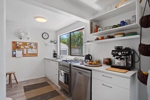 A modern kitchen with white cabinetry, a built-in oven, a dishwasher, and a coffee machine. - The Hideaway @ Whanga (Whangamatā)