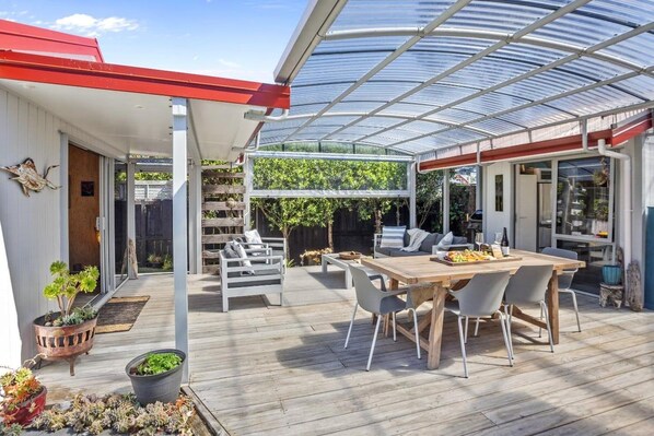Une terrasse extérieure couverte, avec une table et des chaises en bois, entourée de plantes en pot et d’une structure de serre en verre.