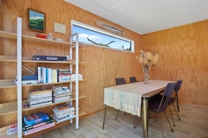 A room with a wooden wall, a bookshelf, a table with chairs, and a window with a view of the sky. - The Hideaway @ Whanga (Whangamatā)