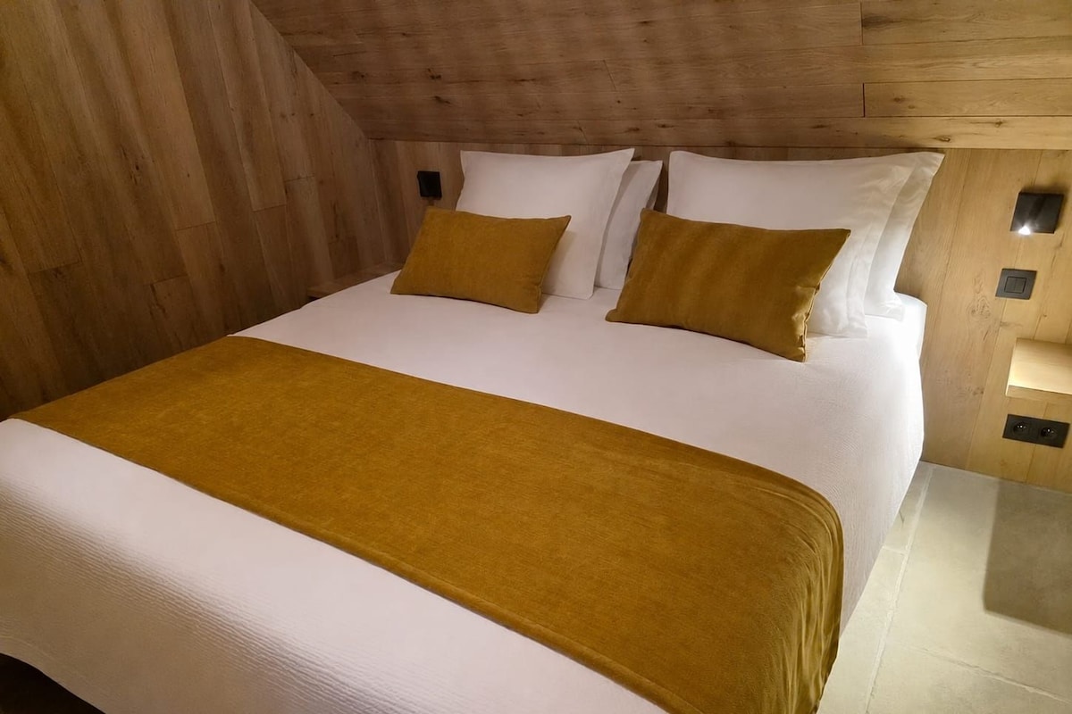 A bedroom with a wooden ceiling, a large bed with white bedding and two mustard-colored pillows, and a bedside table with a lamp.