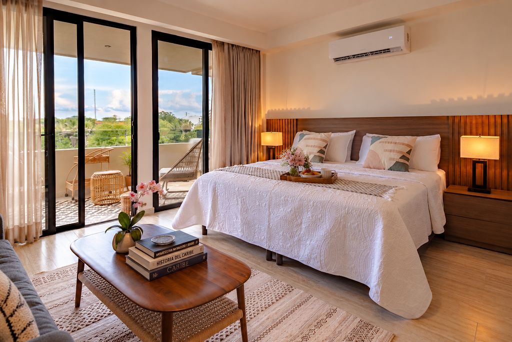 Una habitación de hotel con una cama grande, un balcón con vistas, un escritorio con libros y un aire acondicionado de pared.
