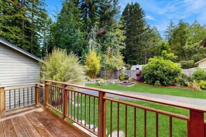 Une terrasse en bois avec une rambarde en métal, une pelouse bien entretenue et divers arbustes et arbres en arrière-plan.