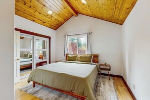 Une chambre avec un plafond en pente, un lit avec un cadre en bois, une table de chevet, une fenêtre avec des rideaux et une porte donnant sur une autre pièce.