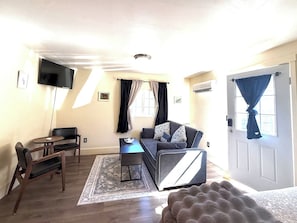 A compact living space with a sofa, a small table, a flat-screen TV mounted on the wall, and a door with curtains.