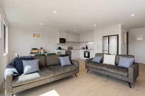 A modern living room with two sofas and a dining table, a kitchen with white cabinets and appliances, and a large mirror.