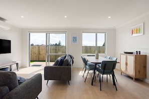A modern living room with a flat-screen TV, a sofa, a dining table with chairs, and a wooden cabinet.