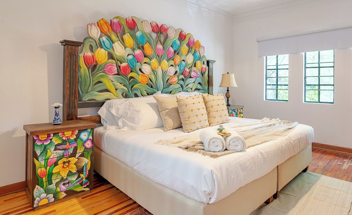 A bedroom with a large bed, a nightstand with a floral design, a window with a view, and a wall-mounted artwork.
