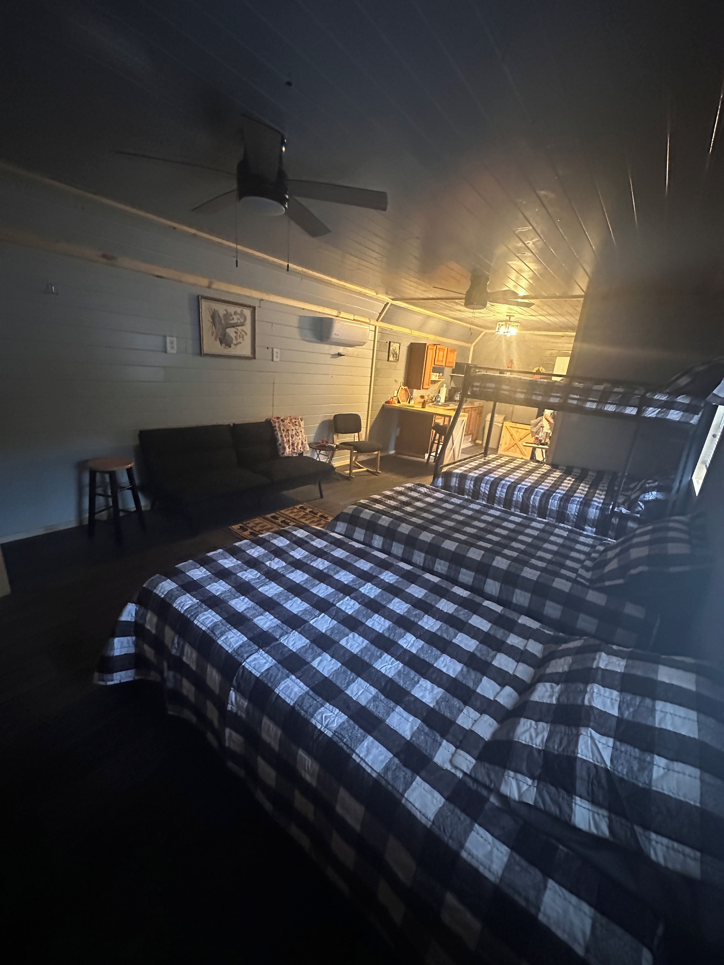 A room with a bed, a chair, a small table, a ceiling fan, and a kitchenette in the background.