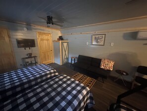 A room with two beds, a ceiling fan, a TV, a wooden door, and a small table with a chair.