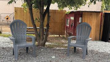 Two Adirondack chairs on a gravel surface with a wooden fence and a red building in the background.