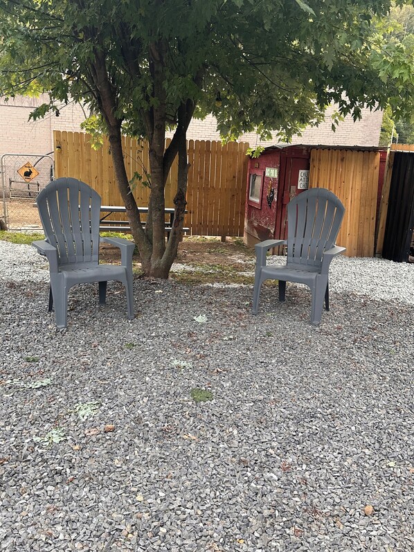 Two Adirondack chairs on a gravel surface with a wooden fence and a red building in the background.