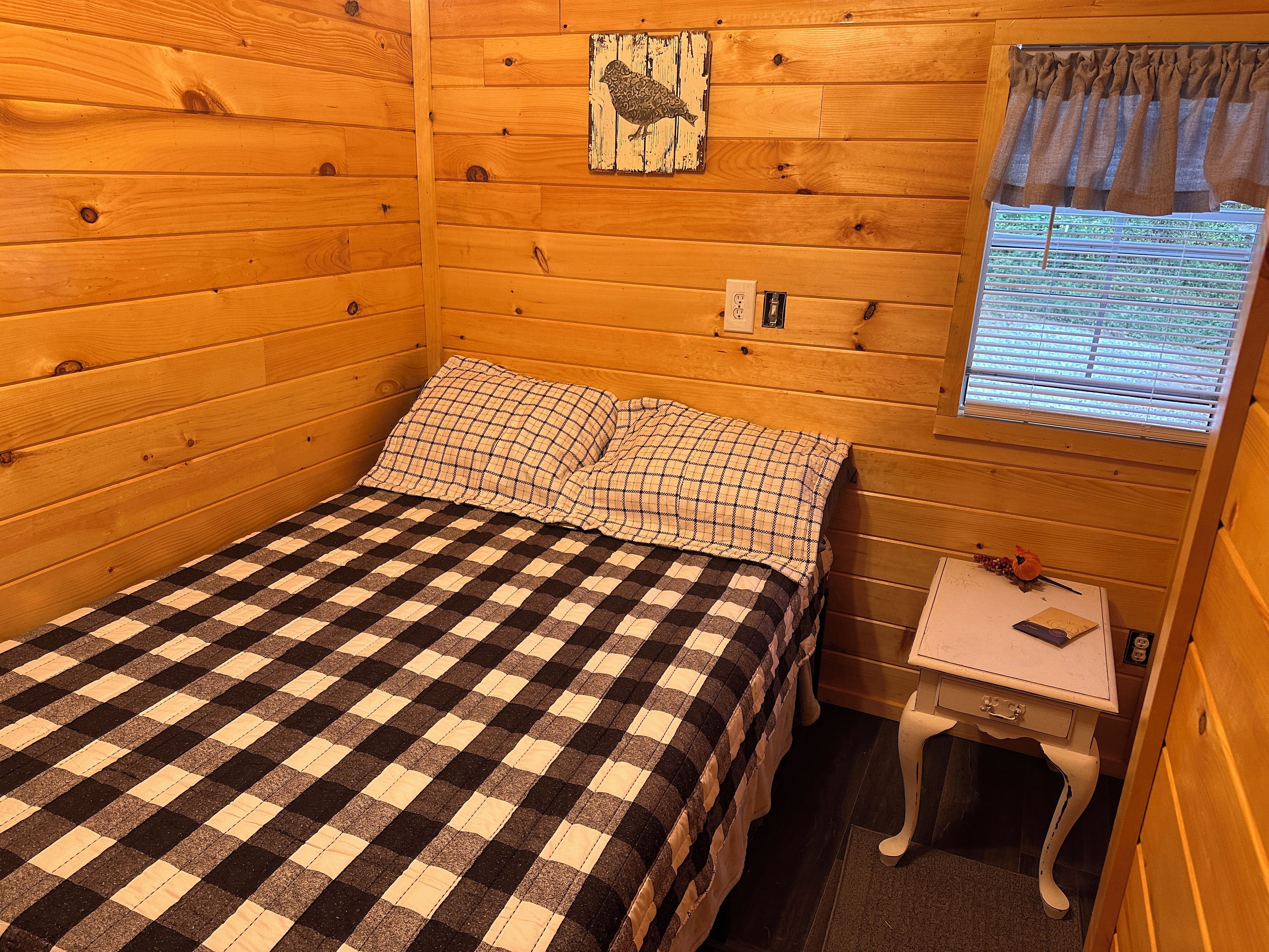 Quarto de cabana de madeira com colcha xadrez, uma pequena mesa com abajur e janela com persianas.