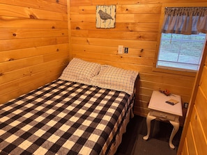 A wooden cabin room with a checkered bedspread, a small table with a lamp, and a window with blinds.