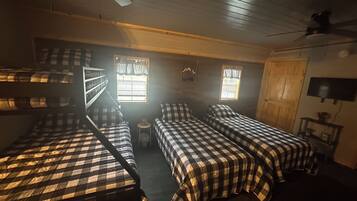 A room with bunk beds, two windows with curtains, a ceiling fan, and a television on the wall.