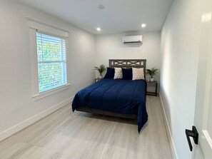 A bedroom with a bed, a window, an air conditioning unit, and a nightstand with a plant. - East Downtown Loft + Full Kitchen (Houston)