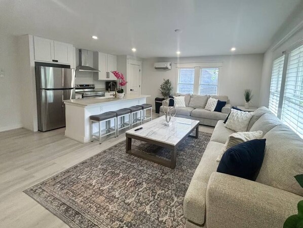 A modern living room with a sofa, coffee table, and a kitchen area with white cabinets and stainless steel appliances. - East Downtown Loft + Full Kitchen (Houston)