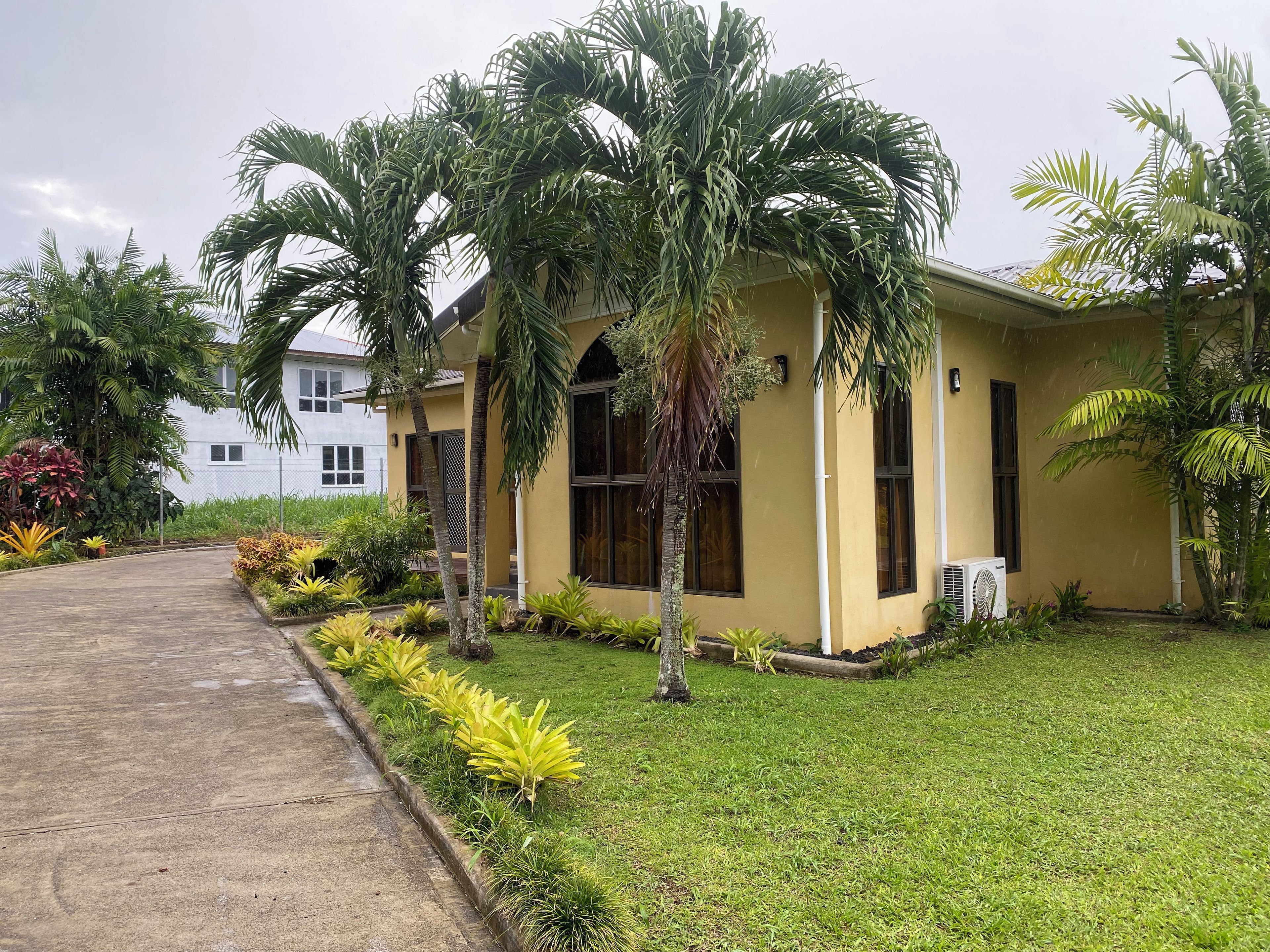 A building with a yellow exterior, surrounded by a well-maintained lawn and various tropical plants.