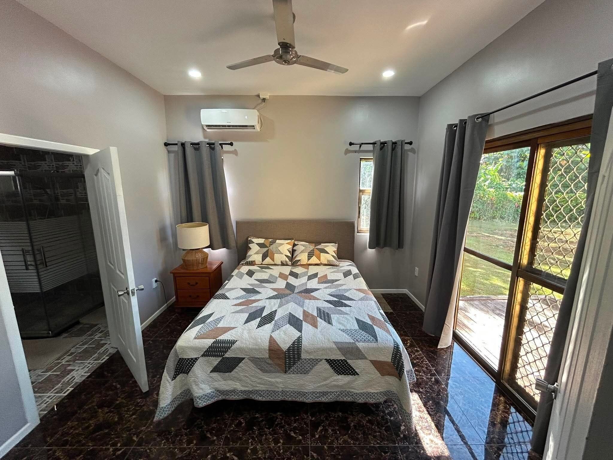 A bedroom with a bed, a ceiling fan, an air conditioning unit, a nightstand, a lamp, and a window with curtains.