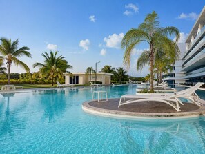 A swimming pool with lounge chairs and palm trees, surrounded by a modern building. - Apartamentoen Cana Bay Punta Cana (Punta Cana)