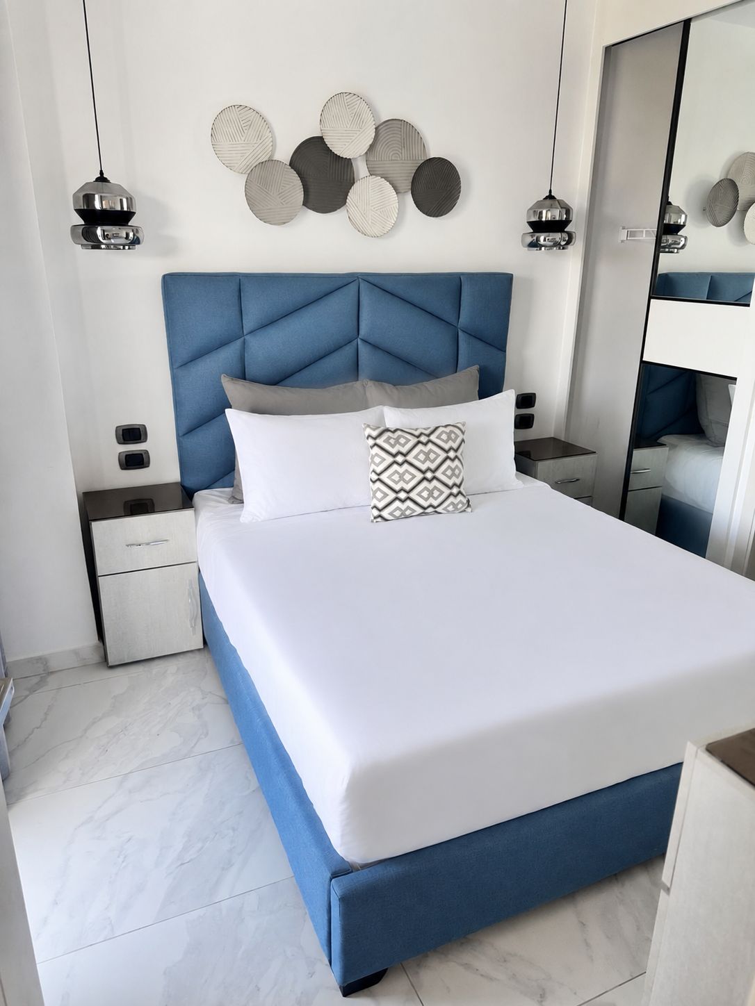 Un dormitorio moderno con cabecera capitonada, una cama grande con ropa de cama blanca, dos mesitas de noche y un espejo.