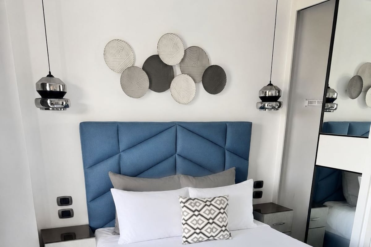 Un dormitorio moderno con cabecera capitonada, una cama grande con ropa de cama blanca, dos mesitas de noche y un espejo.