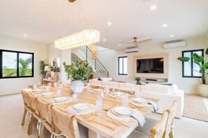 Un comedor moderno con una mesa grande preparada para una comida, un ventilador de techo y un televisor al fondo.