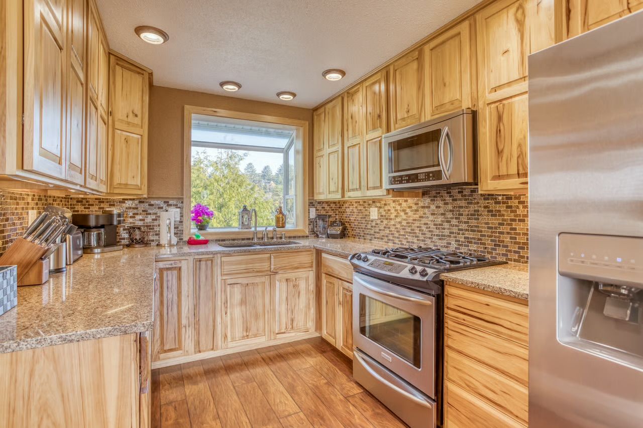 A kitchen with wooden cabinets, a stainless steel refrigerator, a microwave, a stove, and a window with a view of greenery.