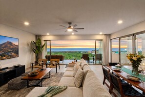 A modern living room with a sofa, chairs, a dining table set for two, and a large window offering a view of a mountainous landscape. - Casa Estrella | 4BD Villa | Private Rooftop Pool (Cabo San Lucas)