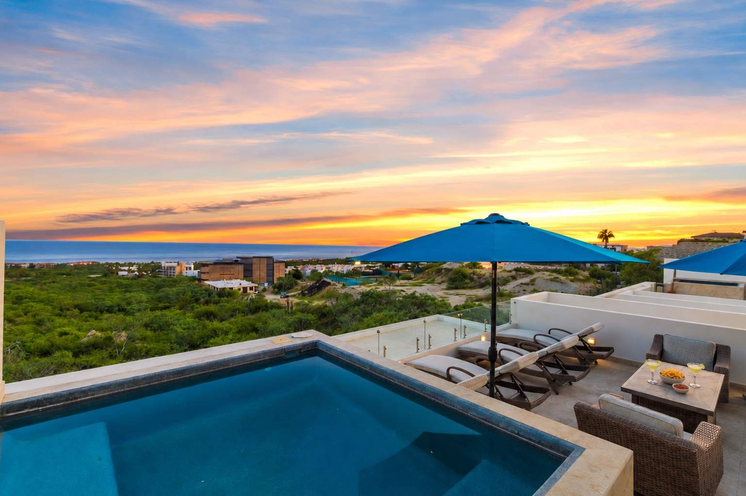 Ein unendlicher Pool mit Liegestühlen und Sonnenschirmen mit Blick auf eine Küstenlandschaft bei Sonnenuntergang.