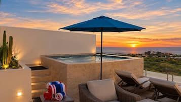 A rooftop terrace with a hot tub, lounge chairs, and an umbrella, overlooking a sunset view.