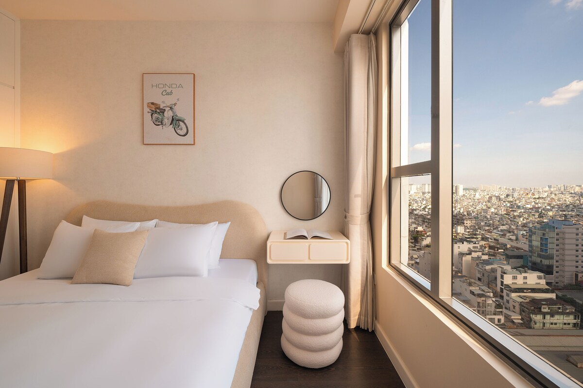 A hotel room with a bed, a bedside table with a mirror, a window with a city view, and a framed picture on the wall.