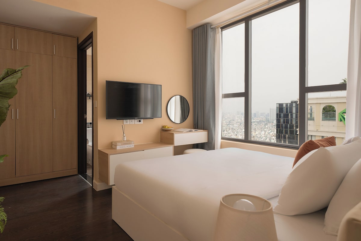 A hotel room with a bed, a TV, a bedside table with a bowl of fruit, a mirror, and a window with a city view.