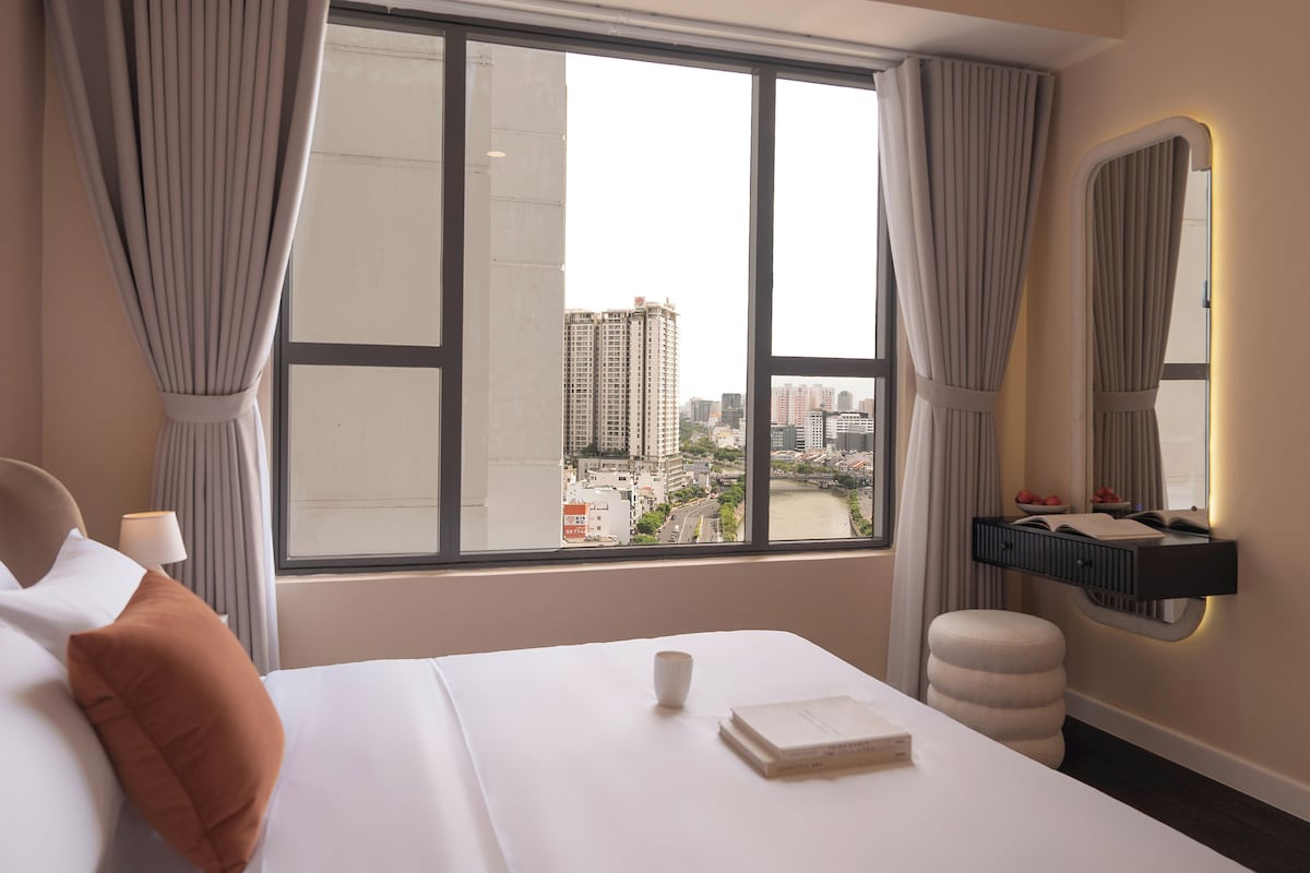 A hotel room with a large window, a bed, a bedside table with a book and a cup, and a mirror on the wall.