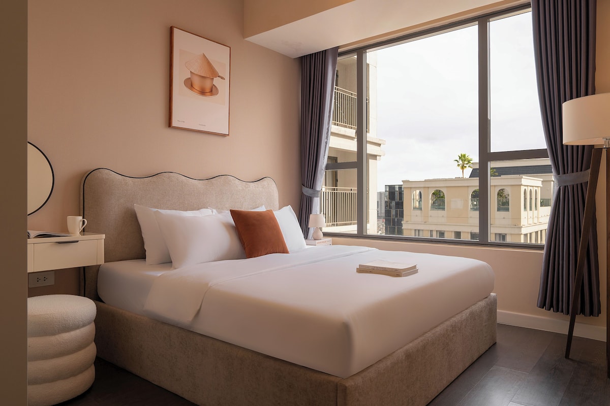 A hotel room with a large bed, a bedside table with a lamp, a window with curtains, and a framed picture on the wall.